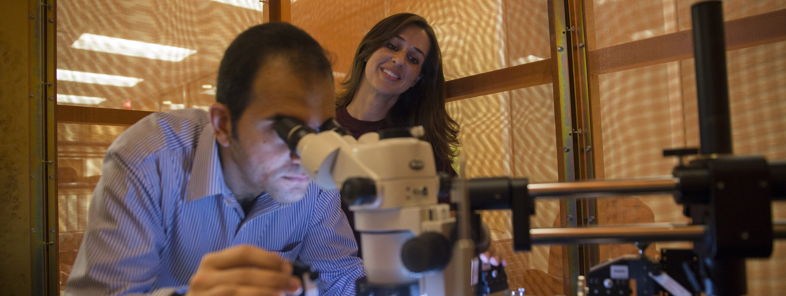 A man looks through a microscope while a woman looks on behind him
