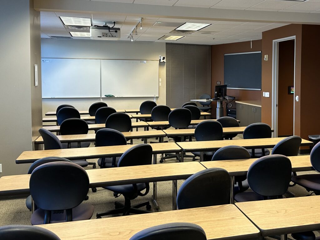 Classroom with rows of tables lined with chairs facing away from the camera. There is an instructor podium in the background in the right hand corner. A whiteboard is hung on the wall and next to it a camera. There is a door to the hallway on the left.